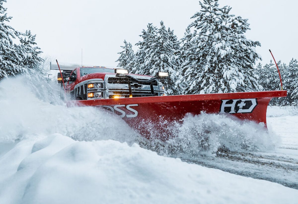 Red BOSS HD straight-blade snowplow on a pickup truck pushing deep snow past snow-covered pine trees