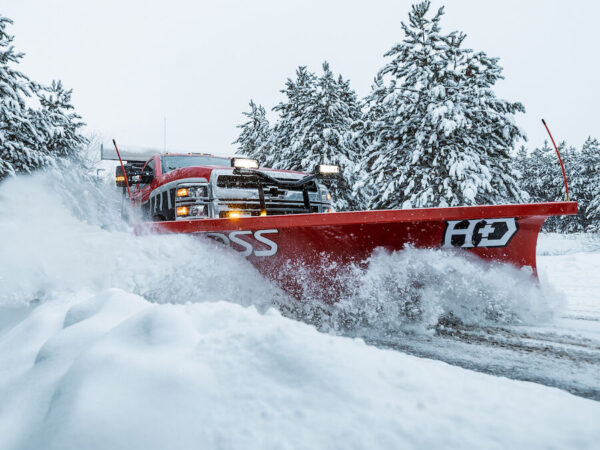 BOSS HD Snowplow Red BOSS HD straight-blade snowplow on a pickup truck pushing deep snow past snow-covered pine trees