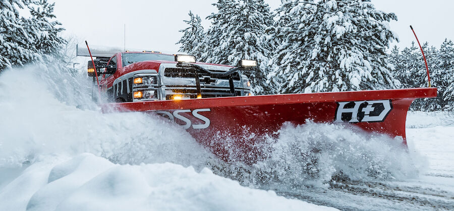 Red BOSS HD straight-blade snowplow on a pickup truck pushing deep snow past snow-covered pine trees