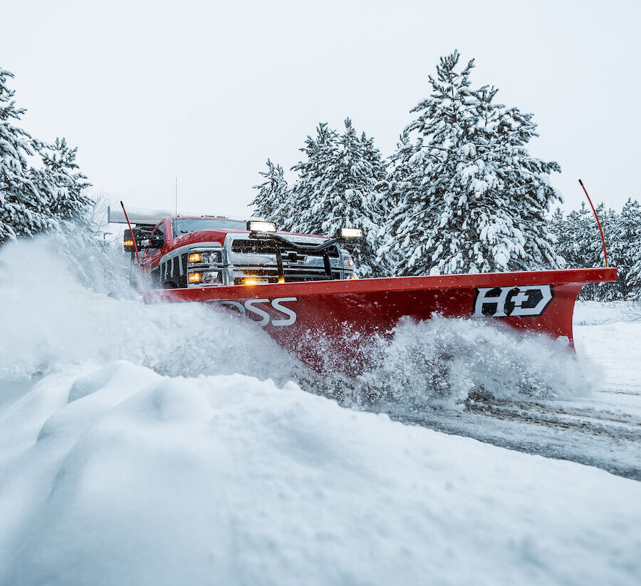 Red BOSS HD straight-blade snowplow on a pickup truck pushing deep snow past snow-covered pine trees