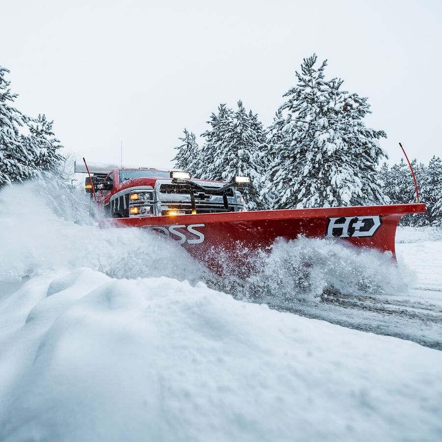 Red BOSS HD straight-blade snowplow on a pickup truck pushing deep snow past snow-covered pine trees