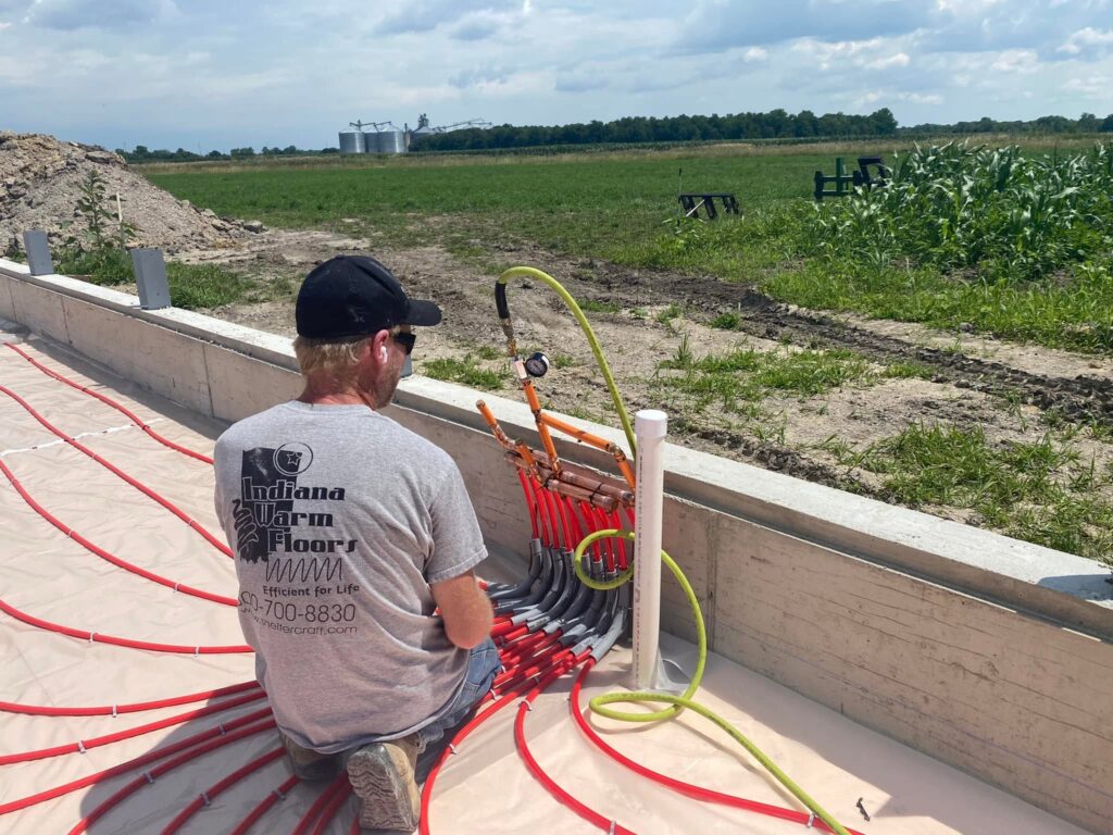 Indiana Warm Floors technician pressure-testing a radiant heating manifold in a farm building beside a cornfield